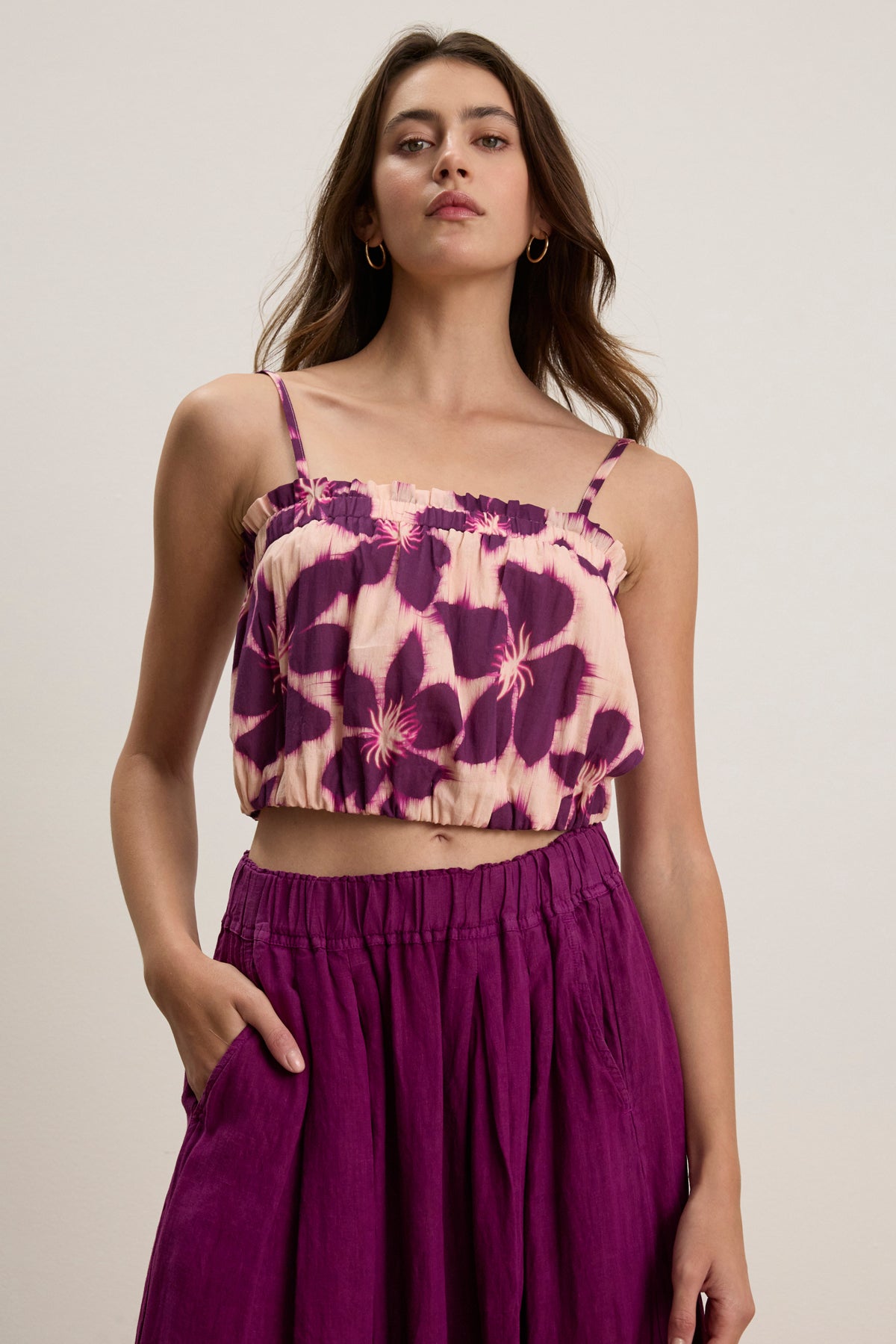 A woman poses against a plain background wearing the ALICE CAMI by Velvet by Graham & Spencer, featuring adjustable straps and a purple and cream floral print, paired with a matching high-waisted purple skirt, one hand in her pocket.