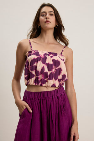 A woman poses against a plain background wearing the ALICE CAMI by Velvet by Graham & Spencer, featuring adjustable straps and a purple and cream floral print, paired with a matching high-waisted purple skirt, one hand in her pocket.