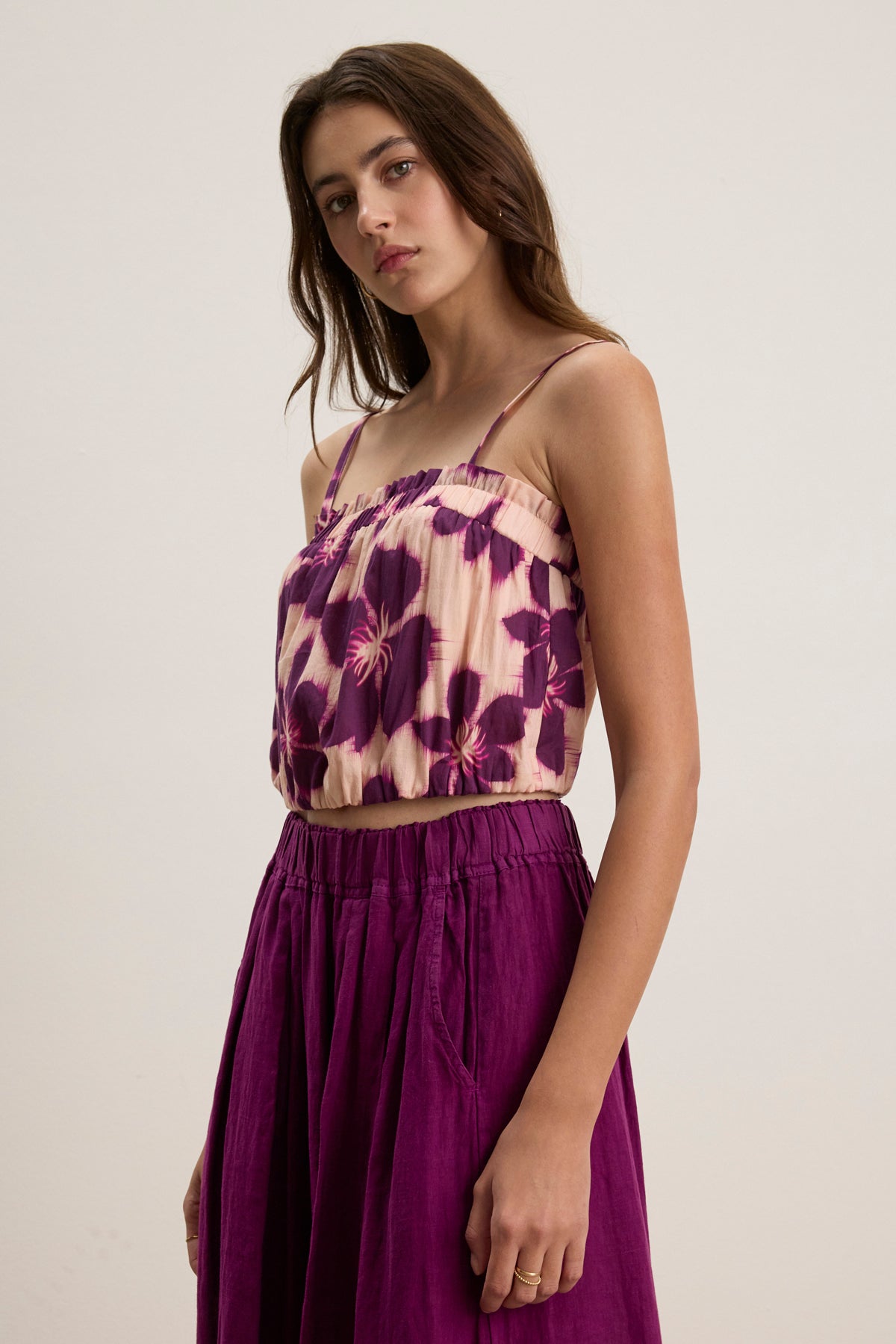 A woman wears the ALICE CAMI by Velvet by Graham & Spencer, a sleeveless, purple floral top with adjustable straps, paired with a matching high-waisted skirt against a plain background.-39111850655937