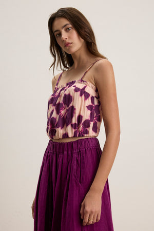 A woman wears the ALICE CAMI by Velvet by Graham & Spencer, a sleeveless, purple floral top with adjustable straps, paired with a matching high-waisted skirt against a plain background.