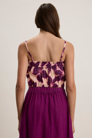 A woman stands facing away, wearing the ALICE CAMI by Velvet by Graham & Spencer—a purple printed voile cami with adjustable straps—and a matching high-waisted skirt.