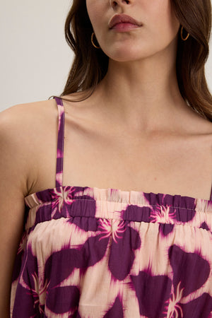 A woman shown from the shoulders up wears the ALICE CAMI, a purple and beige floral-printed voile dress with adjustable straps by Velvet by Graham & Spencer, paired with gold hoop earrings.