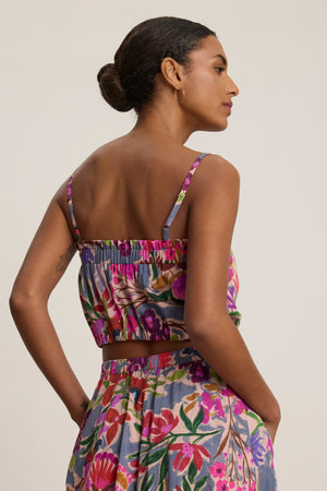 A woman stands sideways with her hands in her pockets, wearing the Velvet by Graham & Spencer ALICE CAMI, a matching floral cropped cami with adjustable straps and skirt set made of printed voile, against a plain background.