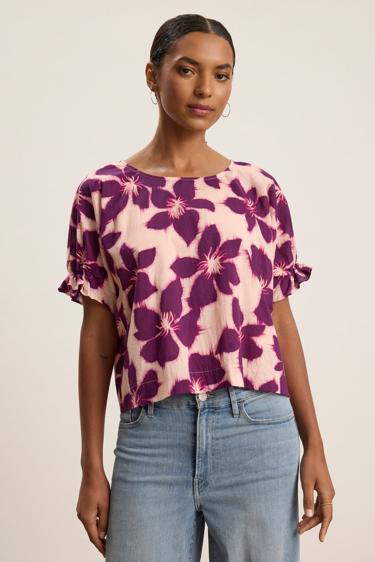 A woman wears the DELILAH BLOUSE by Velvet by Graham & Spencer, a pink cotton voile top with a purple floral print, paired with light blue jeans, standing against a plain light background.