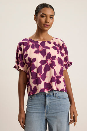 A woman wears the DELILAH BLOUSE by Velvet by Graham & Spencer, a pink cotton voile top with a purple floral print, paired with light blue jeans, standing against a plain light background.