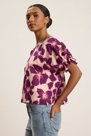 A woman stands sideways in the DELILAH BLOUSE by Velvet by Graham & Spencer— a purple and cream printed, soft cotton voile blouse with dolman sleeves and tie details— paired with light blue jeans against a plain background.
