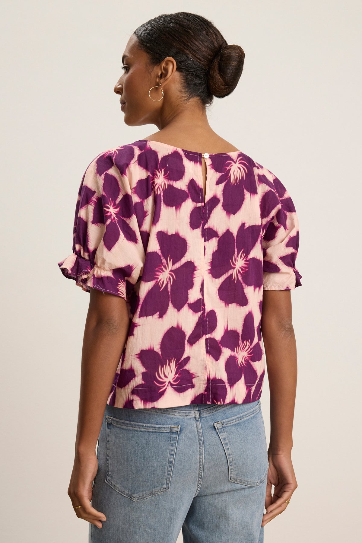 A woman with her hair tied back and gold hoop earrings wears the Velvet by Graham & Spencer DELILAH BLOUSE, a purple and pink floral cotton voile top with dolman sleeves, paired with blue jeans.