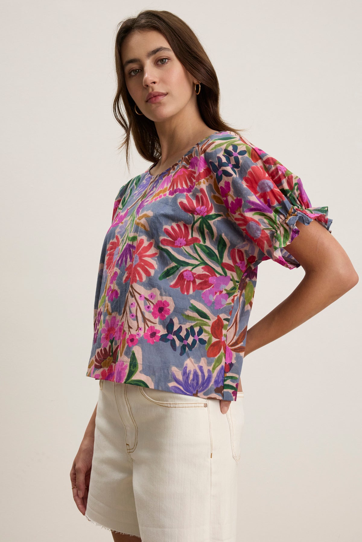 A woman wears the DELILAH BLOUSE by Velvet by Graham & Spencer, featuring a printed design with dolman sleeves in soft cotton voile, paired with cream-colored shorts, standing against a plain light background.