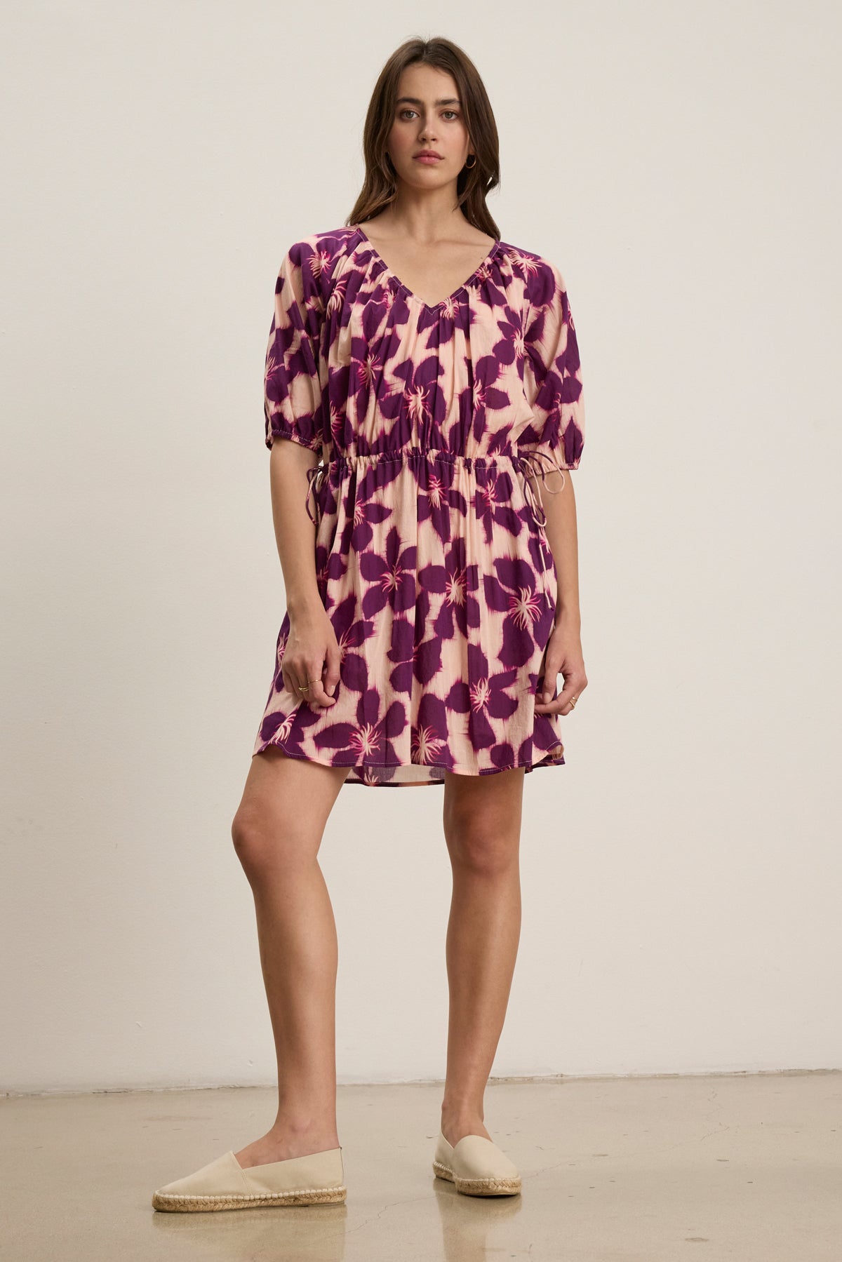 A woman stands indoors wearing the HAZEL DRESS by Velvet by Graham & Spencer, a printed voile dress in purple and beige with an elastic waist and short puff sleeves, paired with light-colored slip-on shoes.