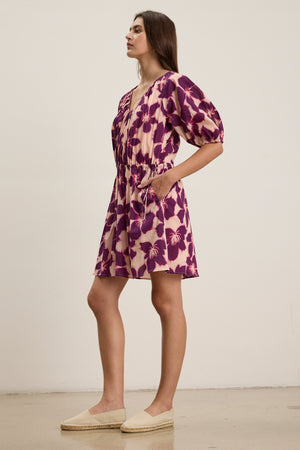 A woman in profile wears the HAZEL DRESS by Velvet by Graham & Spencer, a purple and beige print voile dress with puff sleeves and a drawstring waist, styled with beige espadrilles.
