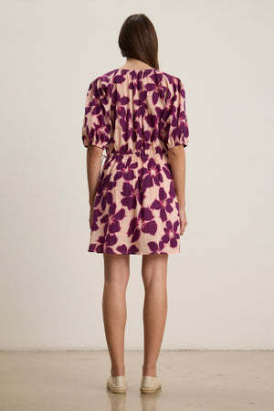 A woman faces away, wearing the HAZEL DRESS by Velvet by Graham & Spencer—a short-sleeved, purple floral printed voile dress—paired with beige espadrilles, set against a plain light background.