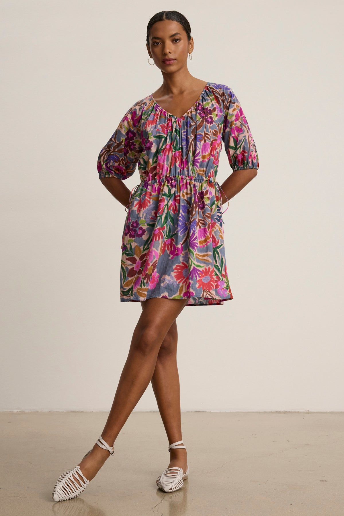 A woman poses against a plain background in the Velvet by Graham & Spencer HAZEL DRESS, a short, colorful printed voile dress with puff sleeves and an elastic waist, paired with white woven flat shoes.-39111852556481