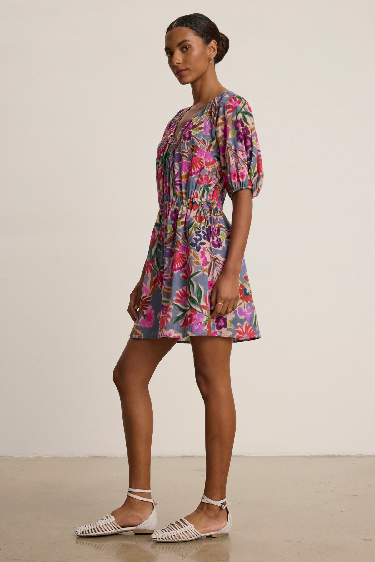 A woman stands sideways in the HAZEL DRESS by Velvet by Graham & Spencer—a short floral dress with puff sleeves and an elastic waist—paired with white woven flats against a plain background.-39111852785857