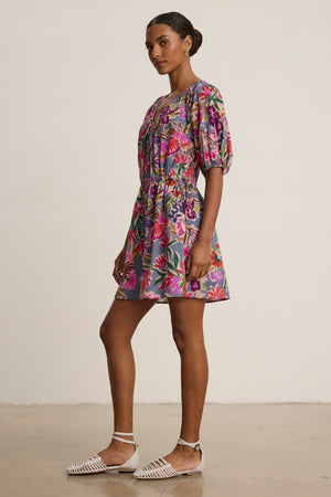 A woman stands sideways in the HAZEL DRESS by Velvet by Graham & Spencer—a short floral dress with puff sleeves and an elastic waist—paired with white woven flats against a plain background.