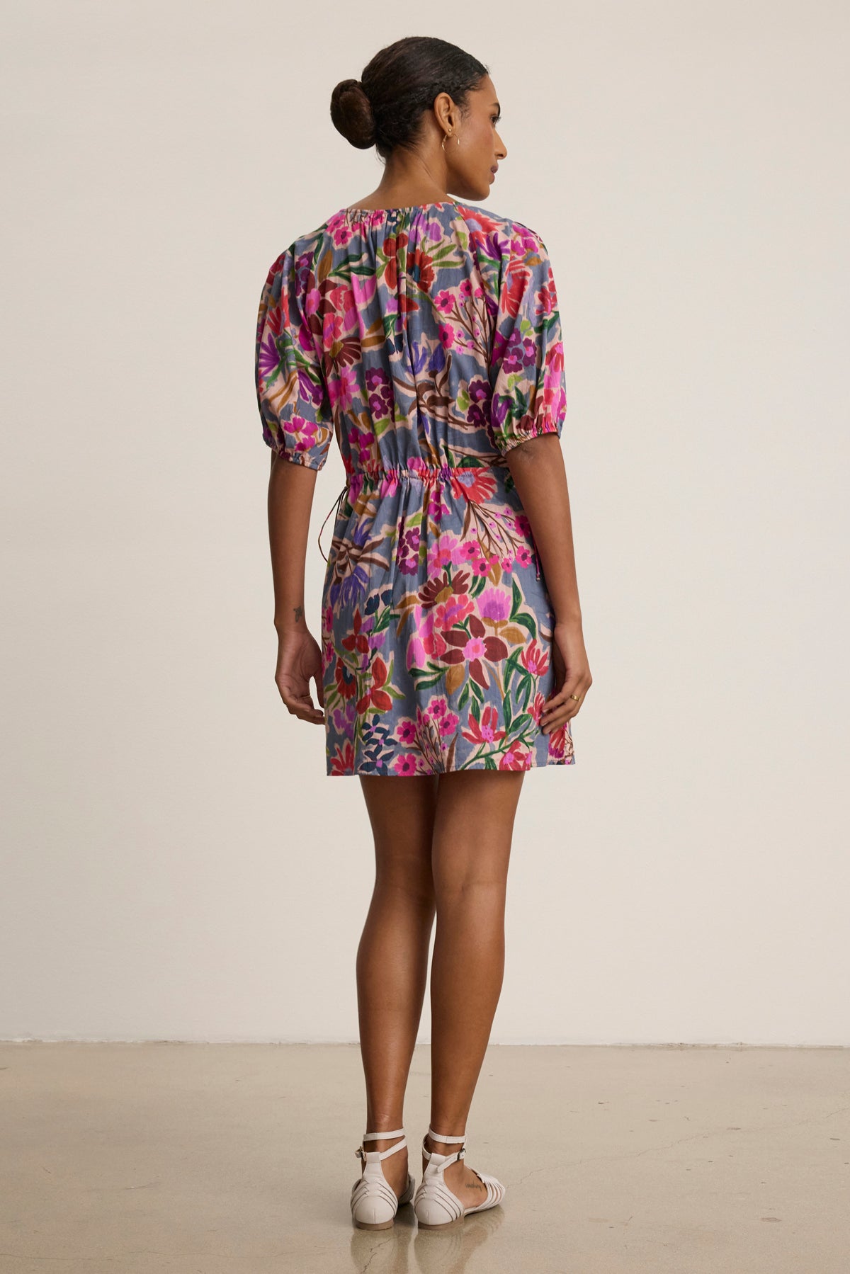 A woman faces away, wearing the HAZEL DRESS by Velvet by Graham & Spencer—a short, colorful floral voile dress with puff sleeves and an elastic waist—paired with white sandals against a plain background.-39111852294337