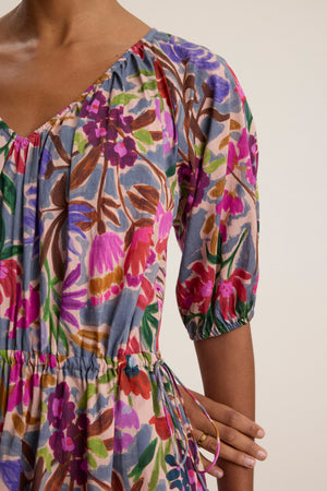 A person wears the HAZEL DRESS by Velvet by Graham & Spencer, a colorful floral-print voile dress with an elastic waist and three-quarter sleeves, shown from neck to mid-thigh.