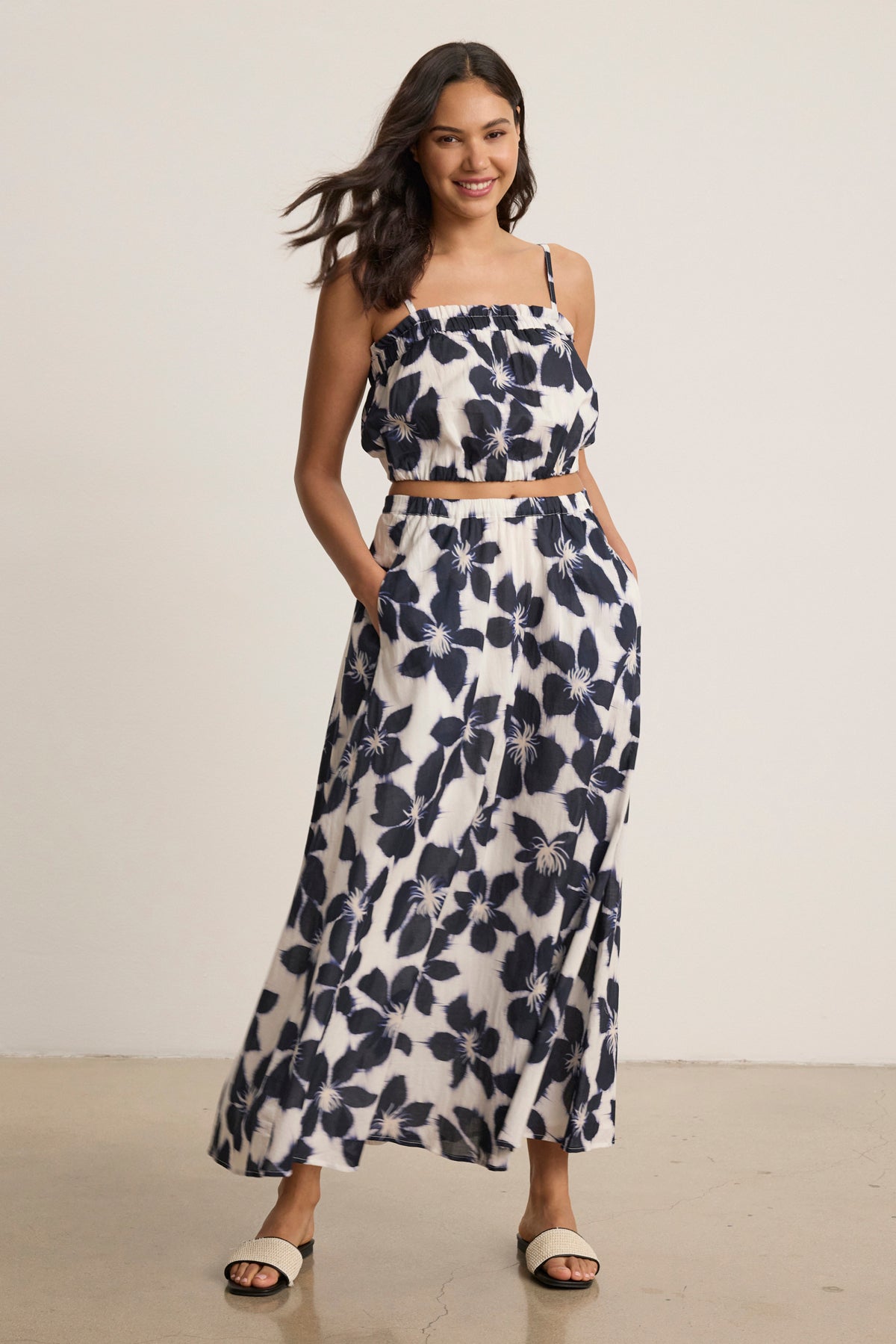 A woman smiles indoors with hands in pockets, showcasing the flowy silhouette of the JESS SKIRT by Velvet by Graham & Spencer—a sleeveless black and white printed voile set with a matching maxi skirt.