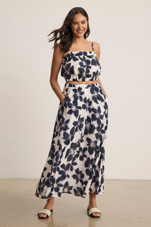 A woman smiles indoors with hands in pockets, showcasing the flowy silhouette of the JESS SKIRT by Velvet by Graham & Spencer—a sleeveless black and white printed voile set with a matching maxi skirt.