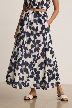 A person wearing the JESS SKIRT by Velvet by Graham & Spencer—a sleeveless crop top and flowy maxi skirt set in a black and white floral print—styled with flat white sandals.