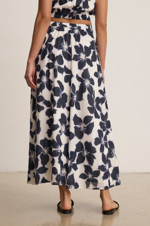 A person wearing the JESS SKIRT by Velvet by Graham & Spencer, featuring a large black floral print, along with a matching cropped top and black sandals, stands on a light-colored floor facing away from the camera.
