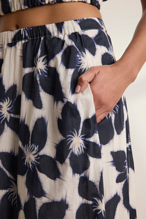 A person stands with one hand in their pocket, wearing the JESS SKIRT by Velvet by Graham & Spencer—a cream voile skirt with bold black floral prints and a flowy silhouette.