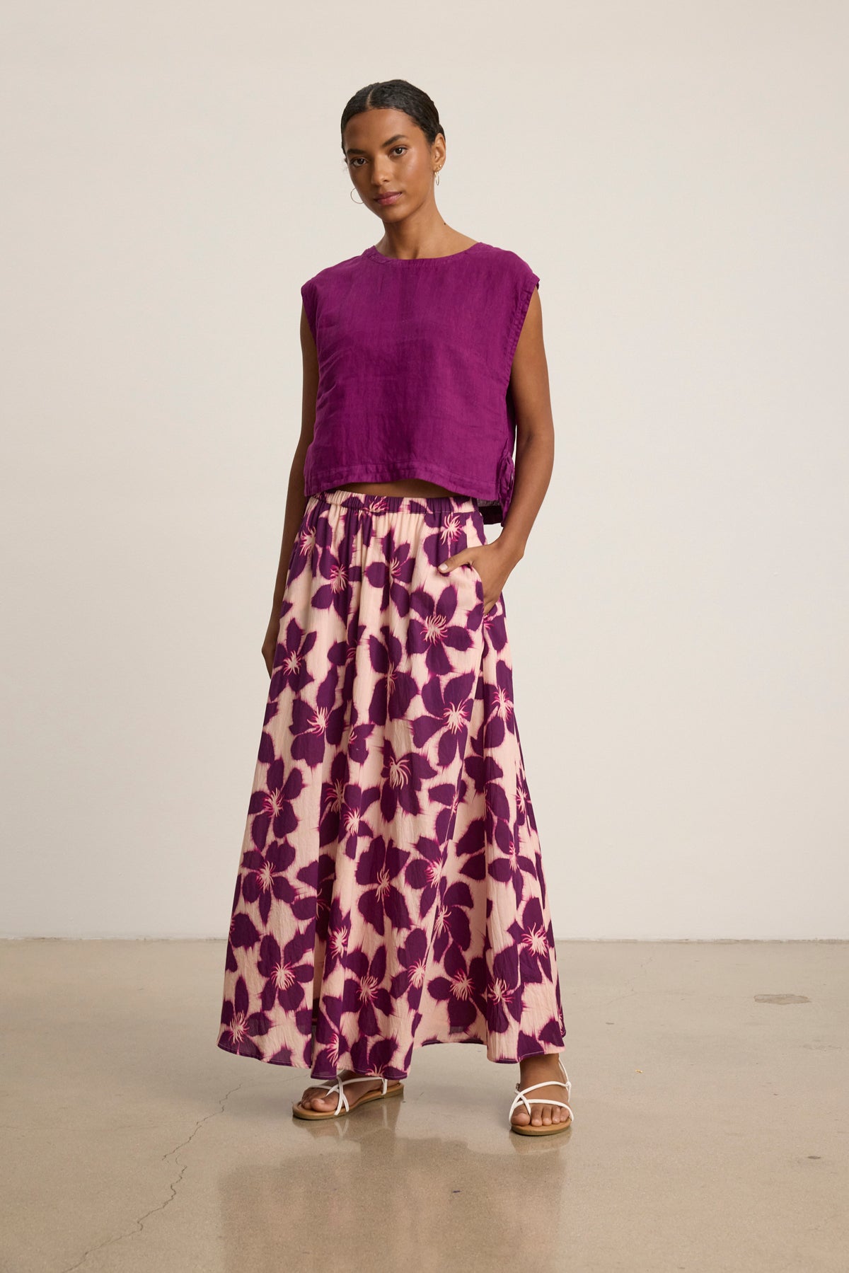 A woman stands indoors wearing a sleeveless purple top and the Velvet by Graham & Spencer JESS SKIRT, a long pink printed voile maxi skirt with a bold purple floral pattern, paired with white sandals.