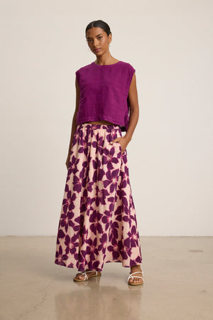 A woman stands indoors wearing a sleeveless purple top and the Velvet by Graham & Spencer JESS SKIRT, a long pink printed voile maxi skirt with a bold purple floral pattern, paired with white sandals.
