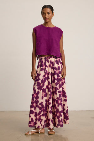 Indoors, a woman wears the Velvet by Graham & Spencer JESS SKIRT—a long, printed voile skirt with bold purple florals—paired with a sleeveless purple top and white sandals for a flowy look.