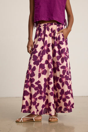 A person wearing the JESS SKIRT by Velvet by Graham & Spencer—a long voile maxi skirt with large purple floral patterns and an elastic waist—pairs it with a sleeveless purple top and white sandals, standing against a plain background.