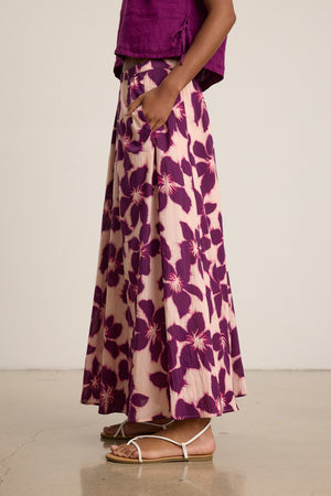 A person wearing the JESS SKIRT by Velvet by Graham & Spencer, a purple floral maxi skirt with an elastic waist, paired with a matching purple top and white flat sandals, stands on a polished floor.