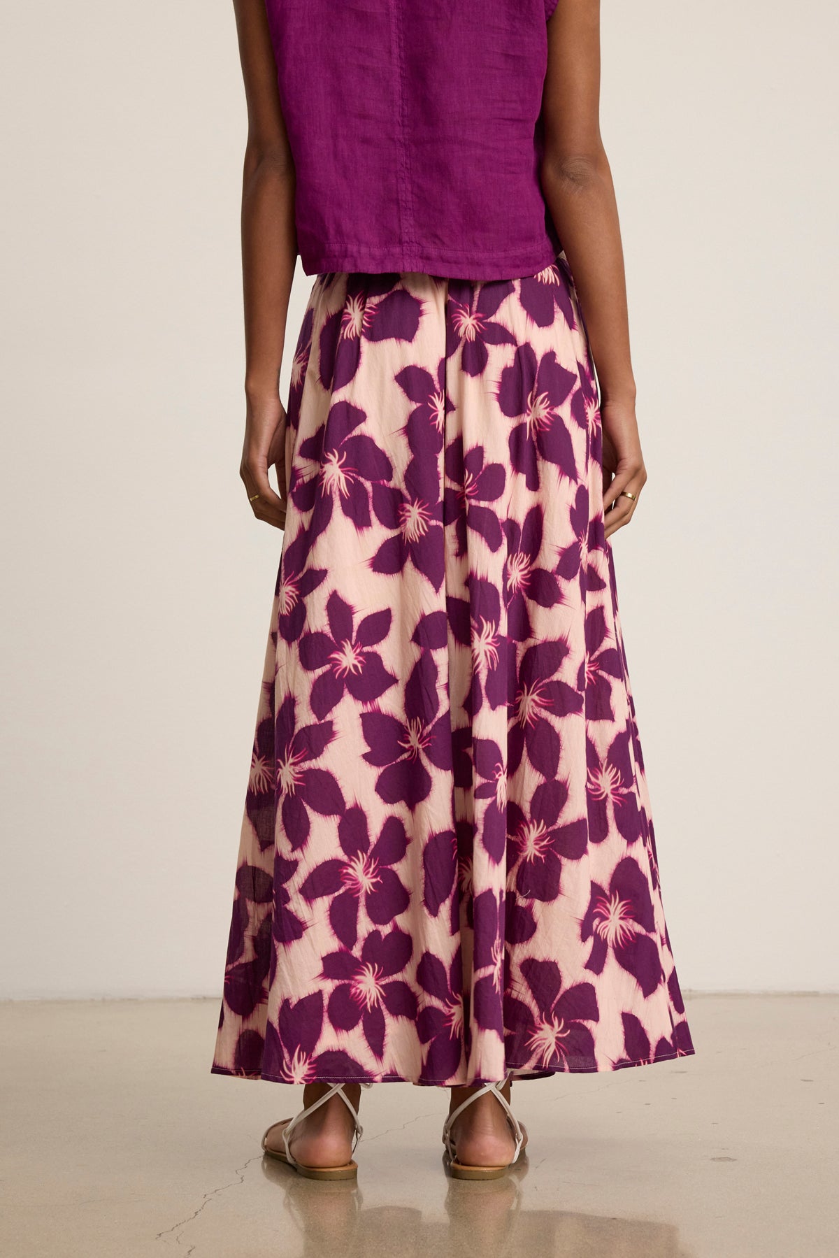 A person stands facing away, wearing a sleeveless purple top and the Velvet by Graham & Spencer JESS SKIRT—a flowy, floral-patterned maxi skirt with an elastic waist—paired with flat sandals.