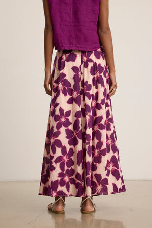 A person stands facing away, wearing a sleeveless purple top and the Velvet by Graham & Spencer JESS SKIRT—a flowy, floral-patterned maxi skirt with an elastic waist—paired with flat sandals.