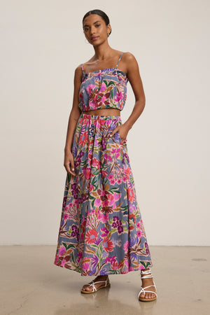 A woman stands in a studio wearing the JESS SKIRT by Velvet by Graham & Spencer, a flowy printed voile maxi skirt in purple, pink, and green tones, styled with a matching floral crop top and white strappy sandals.