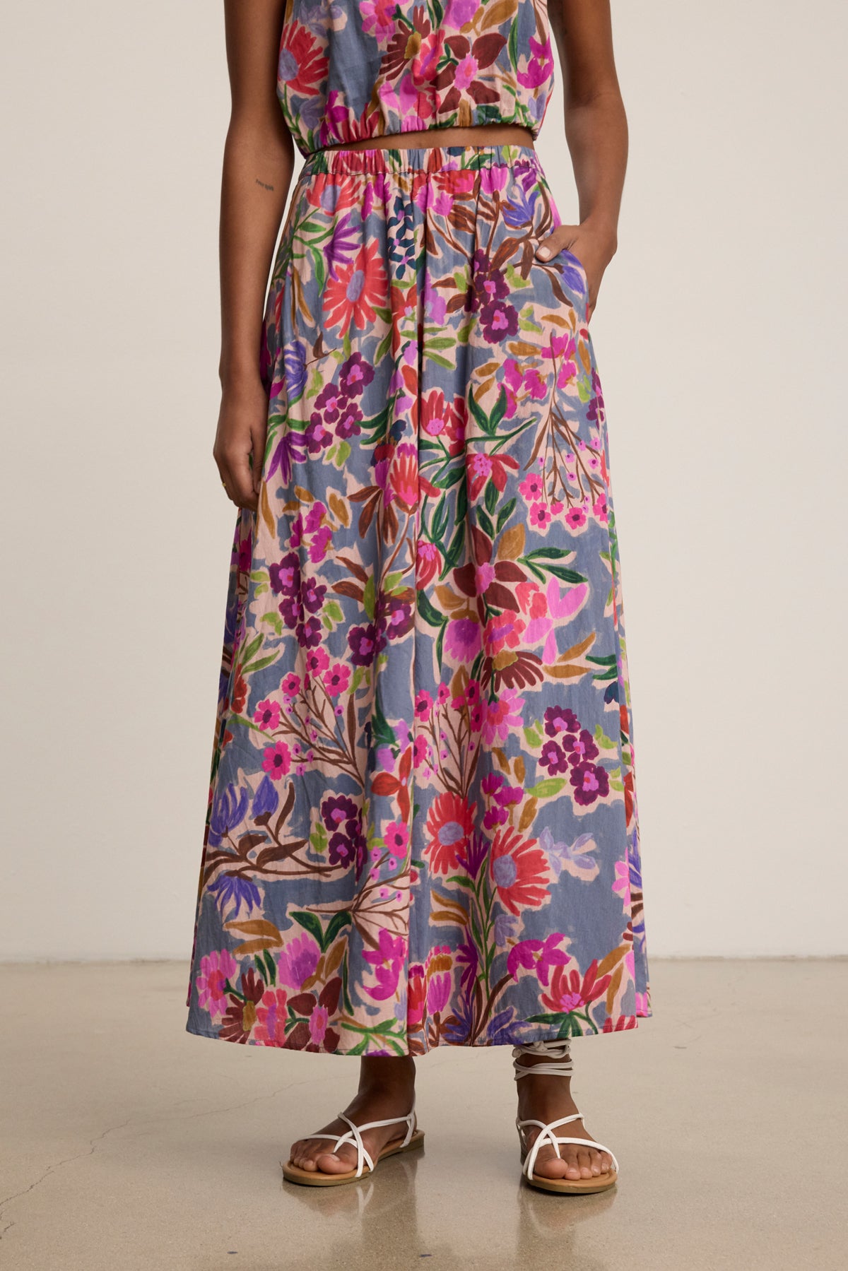 A person wears the JESS SKIRT by Velvet by Graham & Spencer, a printed voile maxi with a flowy silhouette in pink, purple, and green, paired with sandals against a plain background.