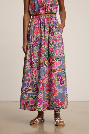 A person wears the JESS SKIRT by Velvet by Graham & Spencer, a printed voile maxi with a flowy silhouette in pink, purple, and green, paired with sandals against a plain background.