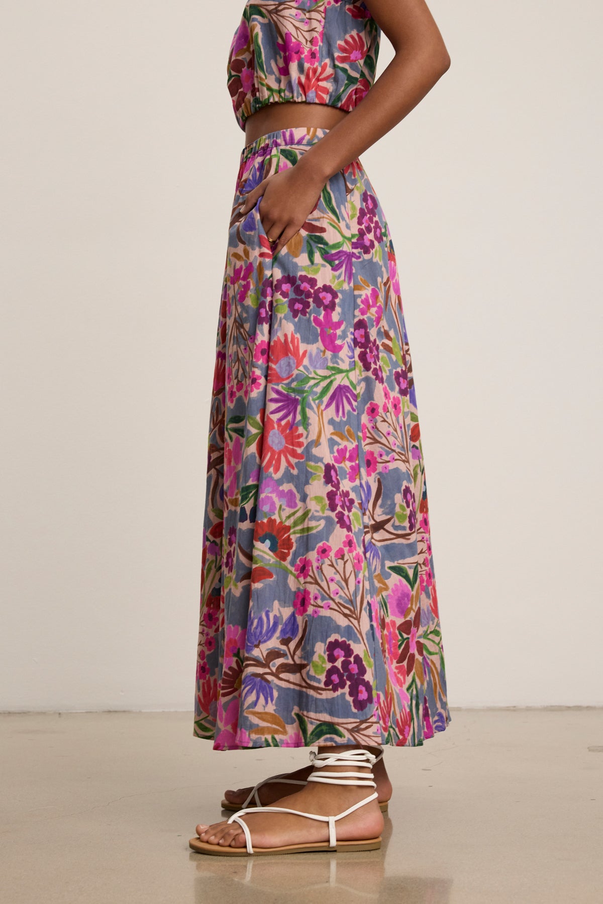 A woman stands sideways against a plain background, wearing the Velvet by Graham & Spencer JESS SKIRT—a colorful printed voile maxi with a flowy silhouette—paired with a matching crop top and white lace-up sandals.