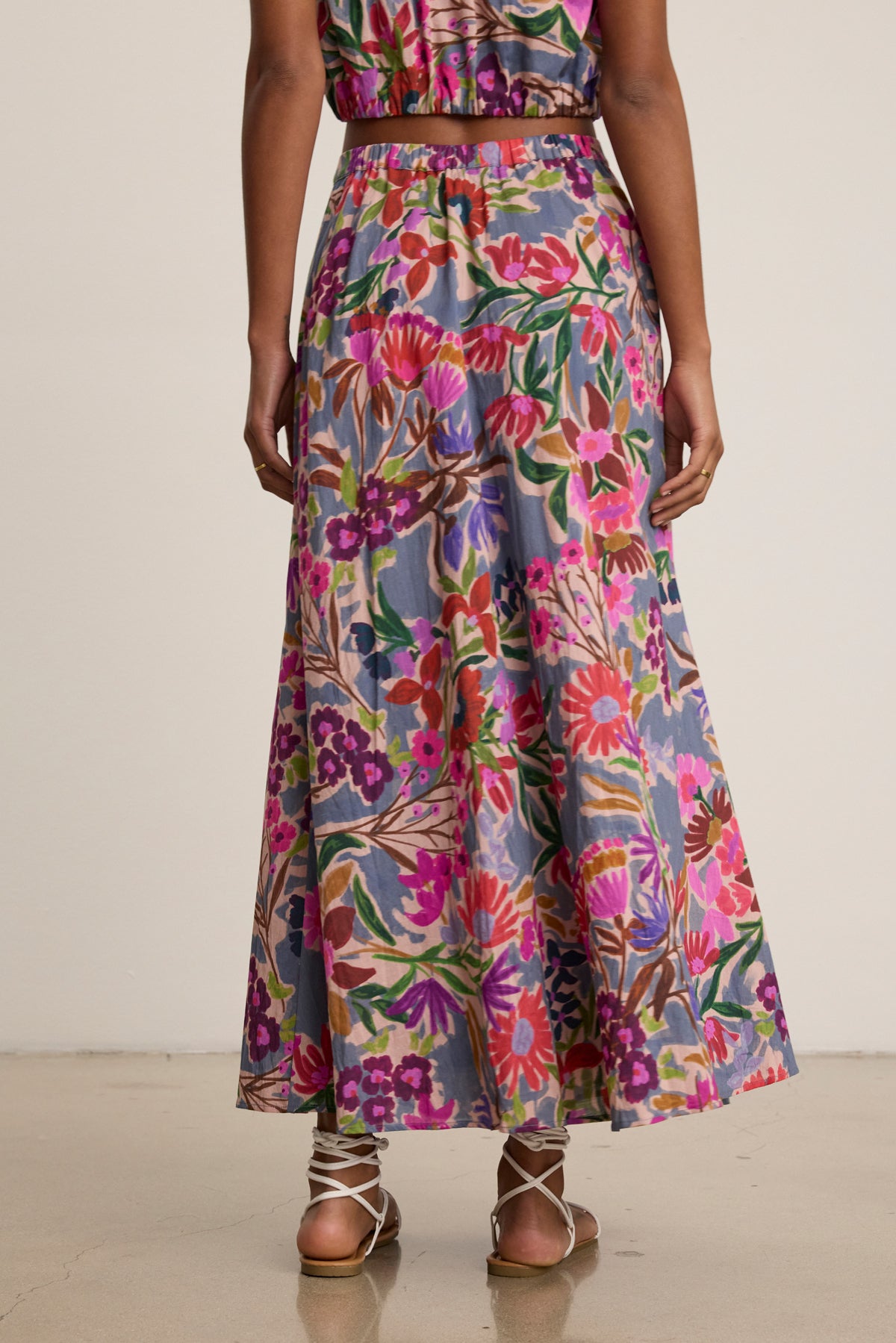 A person stands with their back to the camera, wearing the Velvet by Graham & Spencer JESS SKIRT—a long, printed voile skirt with a flowy silhouette—paired with strappy sandals on a light-colored floor.