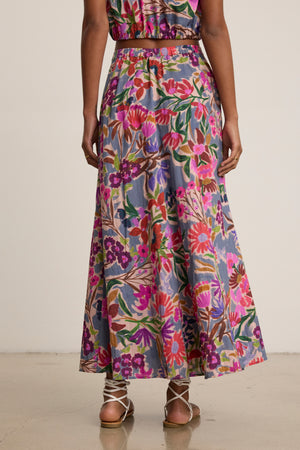 A person stands with their back to the camera, wearing the Velvet by Graham & Spencer JESS SKIRT—a long, printed voile skirt with a flowy silhouette—paired with strappy sandals on a light-colored floor.