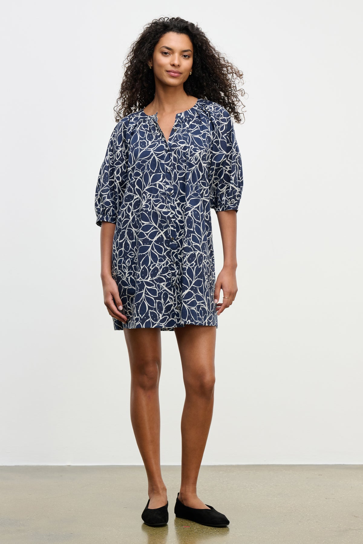 A person wearing the SUMMER DRESS by Velvet by Graham & Spencer—navy blue cotton voile with a white leaf pattern, elbow-length puff sleeves, and black flats—stands against a plain light background.