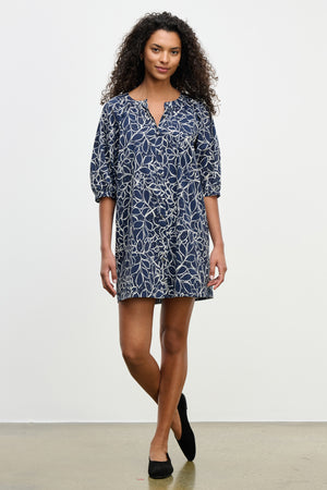 A woman stands against a plain background wearing the SUMMER DRESS by Velvet by Graham & Spencer, a navy cotton voile mini dress with a white floral pattern, puff sleeves, and black flats.