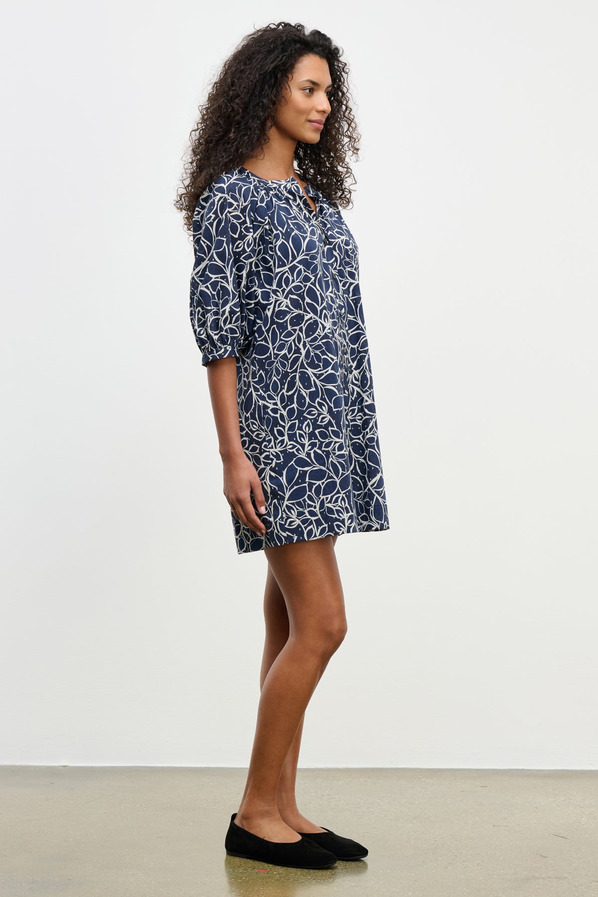 A woman stands sideways against a white background wearing the Velvet by Graham & Spencer SUMMER DRESS—a navy blue, knee-length cotton voile dress with puff sleeves and a white abstract leaf pattern—paired with black flats.