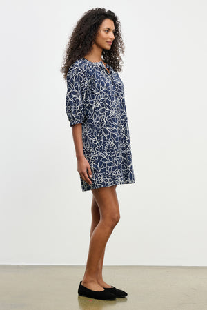 A woman stands sideways against a white background wearing the Velvet by Graham & Spencer SUMMER DRESS—a navy blue, knee-length cotton voile dress with puff sleeves and a white abstract leaf pattern—paired with black flats.