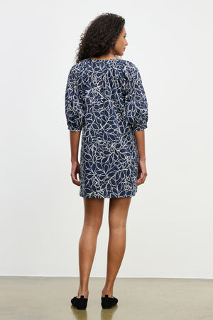 A woman faces away, wearing the SUMMER DRESS by Velvet by Graham & Spencer—navy cotton voile, knee-length, with white abstract leaf prints and elbow-length puff sleeves—set against a plain light background.
