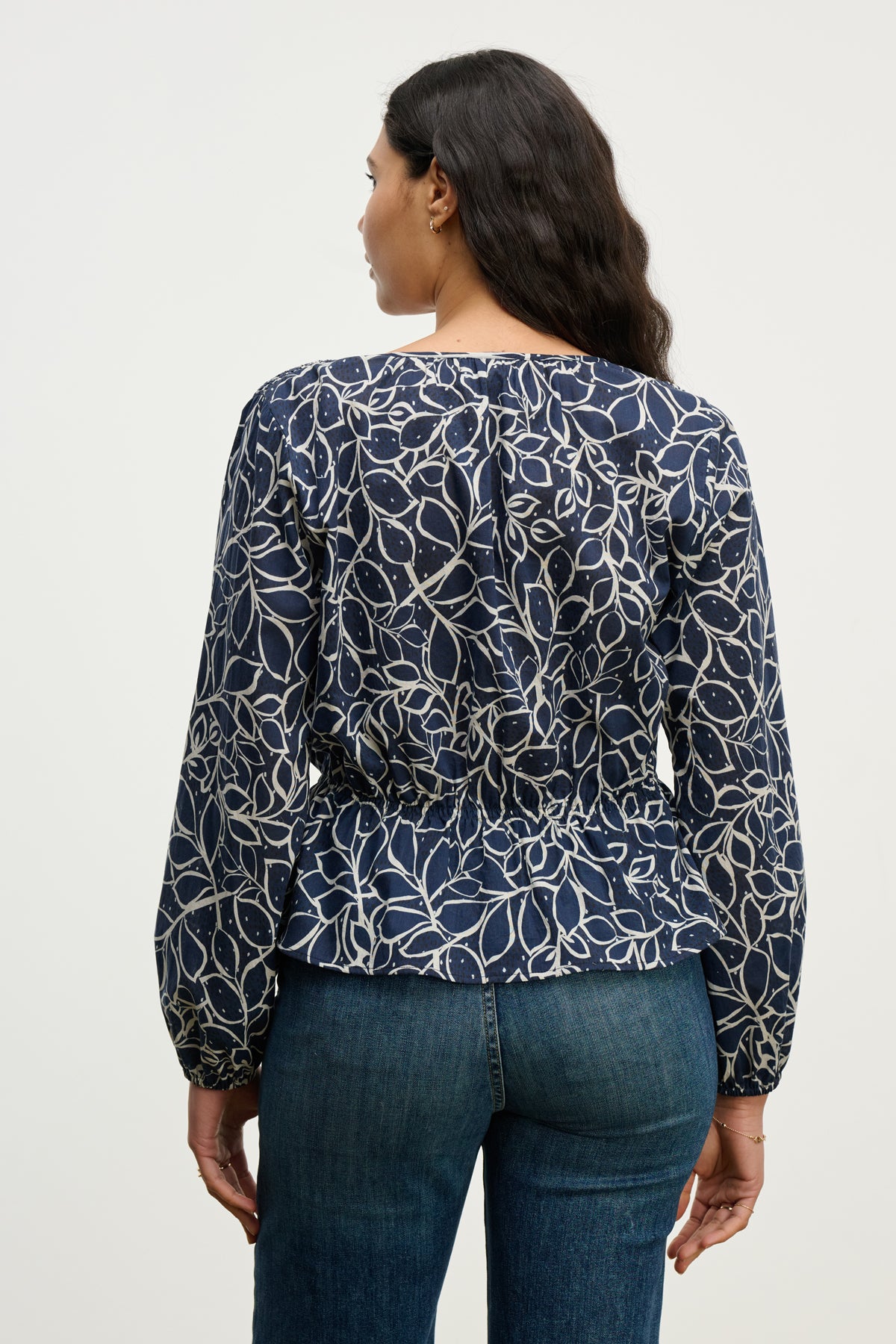 A woman is shown from the back wearing the Velvet by Graham & Spencer WYNNE TOP—a navy cotton voile blouse with a white abstract leaf pattern—paired with blue jeans, highlighting her silhouette against a plain background.-40227172974785