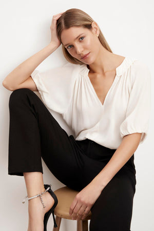 A woman is posing on a stool wearing a Velvet by Graham & Spencer ALEGRA V-NECK TOP with puff sleeves and black pants.