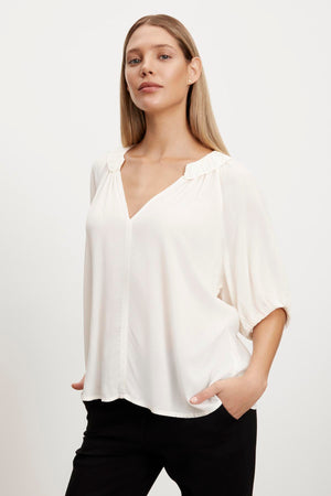 The model is wearing a white ALEGRA V-NECK TOP blouse by Velvet by Graham & Spencer and black pants.
