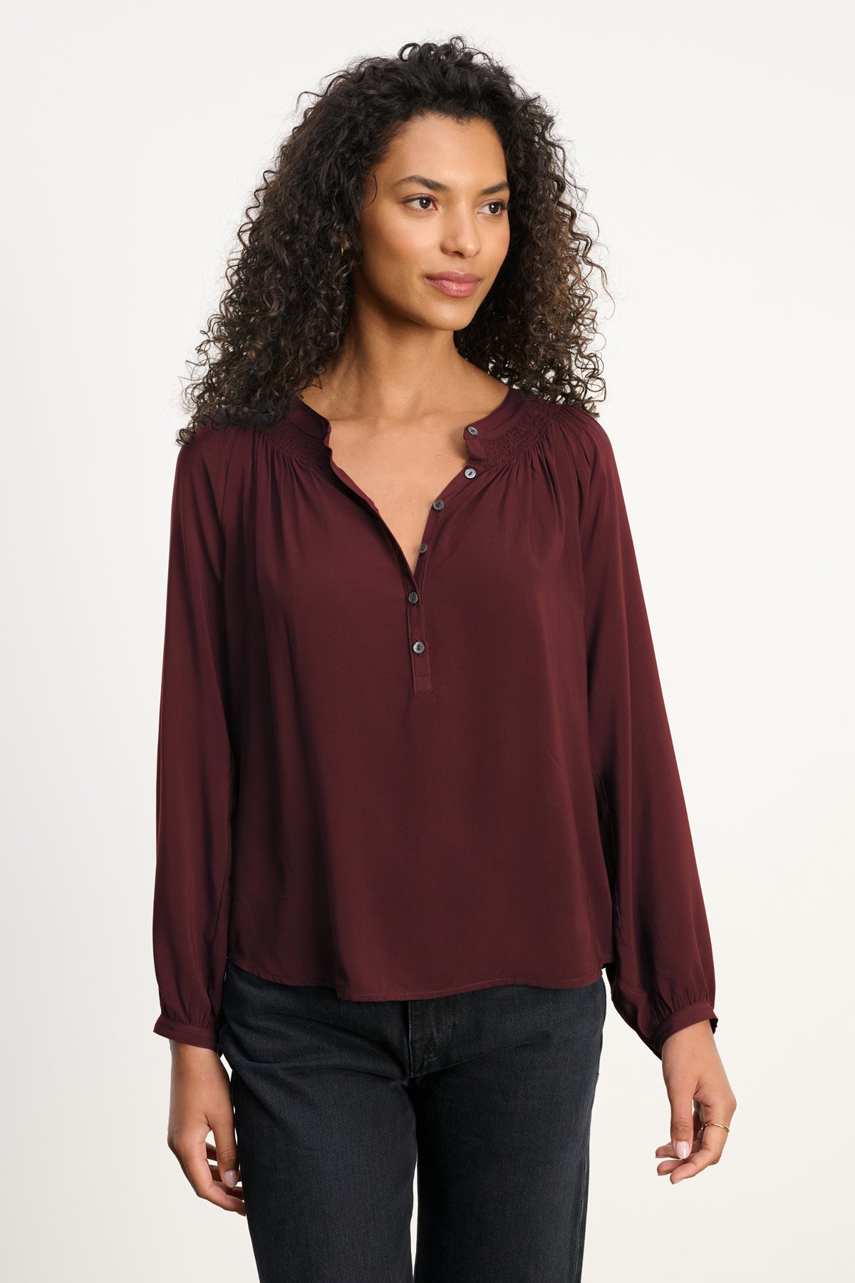 A woman with curly hair wears the AURELIA TOP by Velvet by Graham & Spencer, a burgundy long-sleeve blouse with smocked details, paired with black pants against a plain light background.