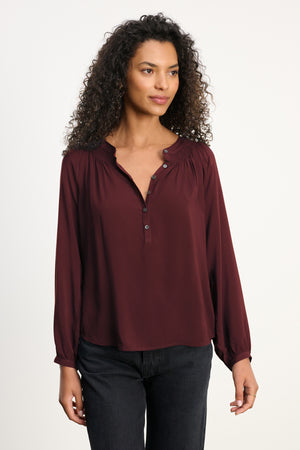 A woman with curly hair wears the AURELIA TOP by Velvet by Graham & Spencer, a burgundy long-sleeve blouse with smocked details, paired with black pants against a plain light background.