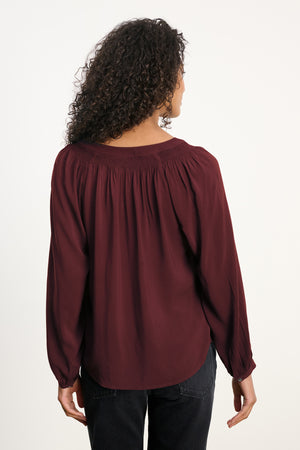 A person with curly hair, shown from the back, wears Velvet by Graham & Spencer's AURELIA TOP—a long-sleeve, burgundy blouse with smocked detailing—paired with black pants.