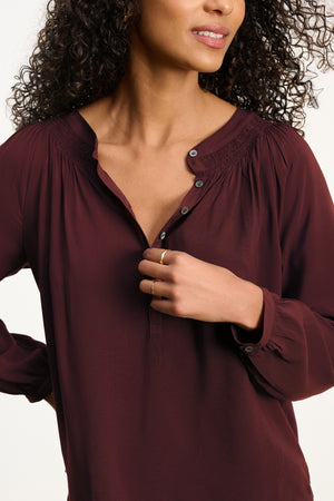 A woman with curly hair, her face partially visible, touches the collar of the maroon AURELIA TOP by Velvet by Graham & Spencer—a relaxed fit, long-sleeve blouse with buttons.